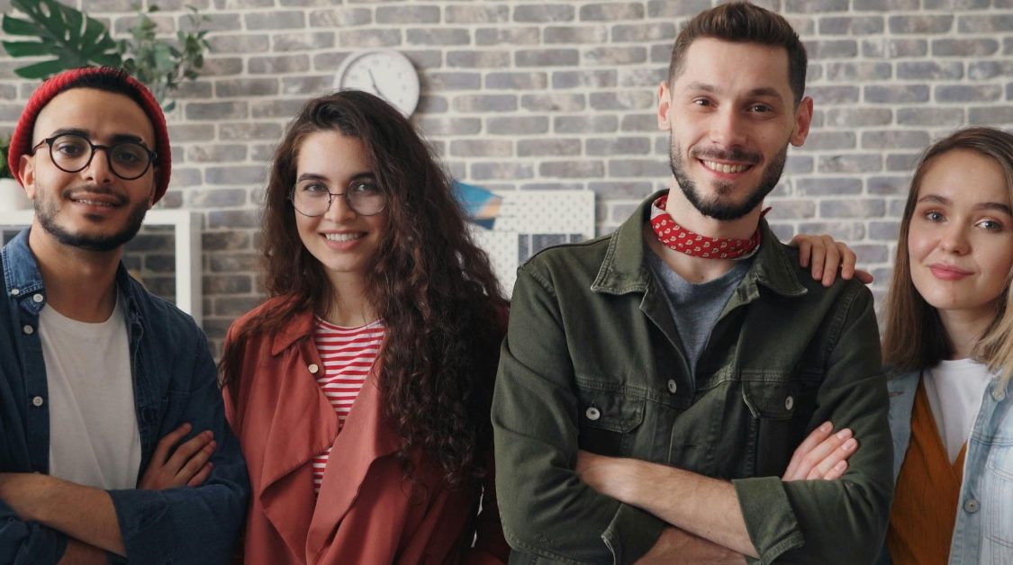 Vier junge Menschen lächeln, stehen arm in arm in einem modernen Raum.