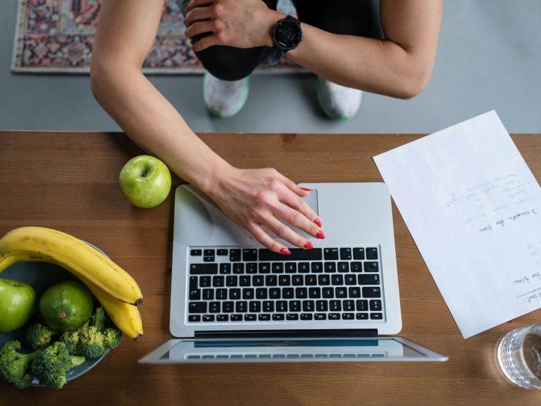 Eine Person arbeitet am Laptop, umgeben von Obst und einem Dokument auf dem Tisch.