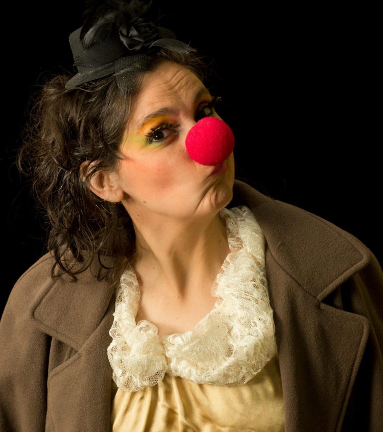 Frau mit roter Nase, bunten Augen und einem Hut, trägt ein Kostüm mit einem auffälligen Kragen.