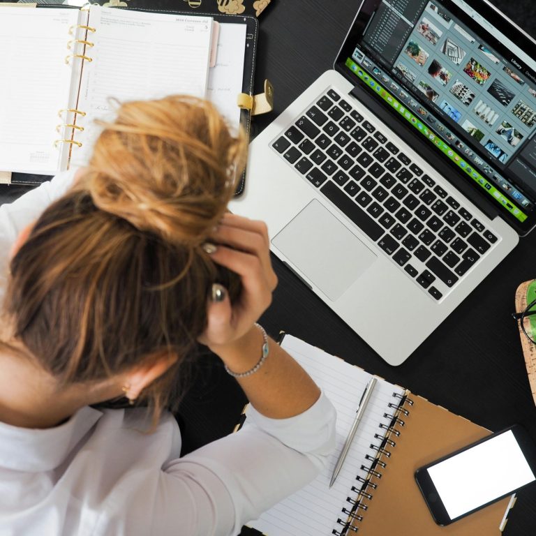 Junge Frau mit gestresstem Gesichtsausdruck sitzt vor Laptop und Notizen.