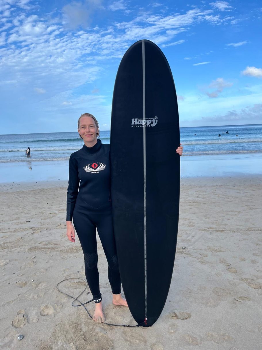 Auch nach Jahren noch Anfängerin. Junge Frau in Neoprenanzug hält ein schwarzes Surfbrett am Strand.