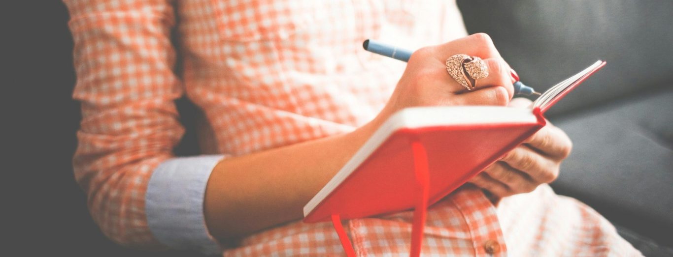 Eine Hand, die in ein rotes Notizbuch schreibt, auf einem grauen Sofa.