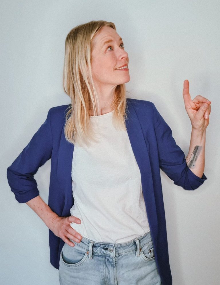 Frau mit blonden Haaren in blauer Jacke, zeigt nach oben und lächelt.