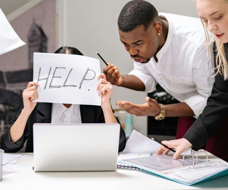 Eine Person hält ein "HELP"-Schild vor einem Laptop, während andere sie ansprechen.