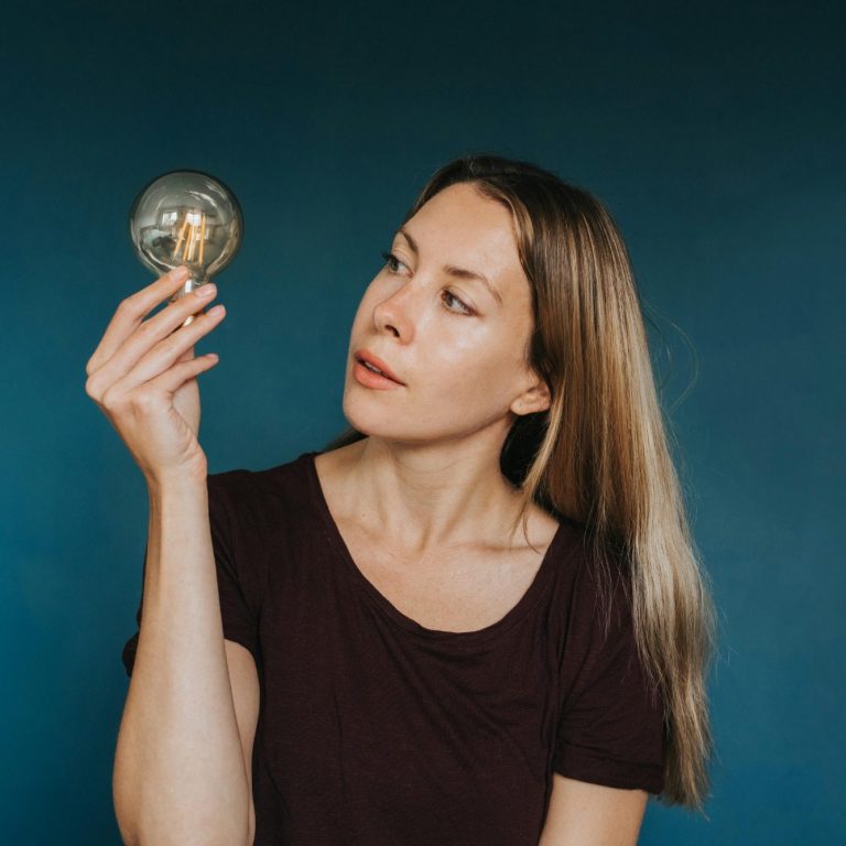 Frau mit langen Haaren hält eine Glühbirne vor einem blauen Hintergrund.