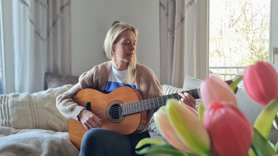 Gitarre spielen entspannt mich. Eine Frau spielt Gitarre auf einem Sofa, umgeben von einem Blumenstrauß.
