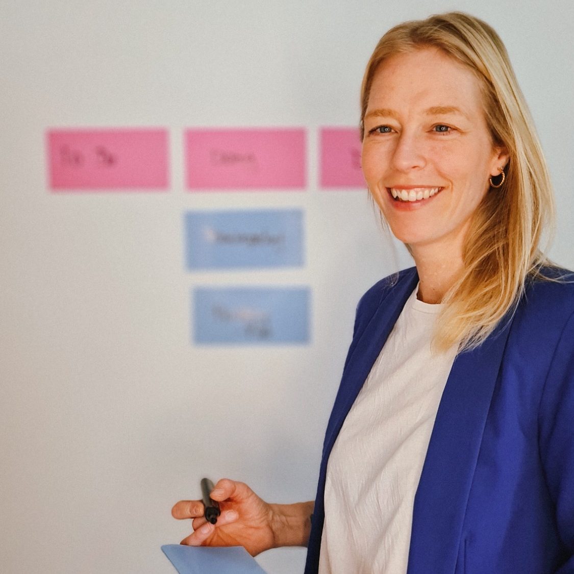 Frau in blauer Jacke lächelt und hält einen Stift vor farbigen Notizen.