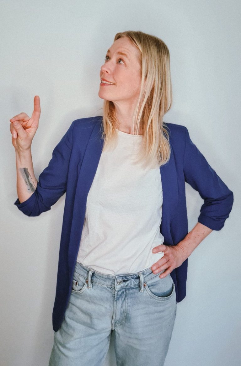 Frau mit blonden Haaren in blauer Jacke hebt eine Hand, schaut nach oben.
