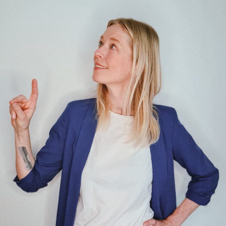 Blonde Frau mit blauen Blazer zeigt nach oben und lächelt.