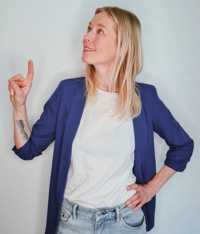 Frau mit blonden Haaren in blauer Jacke zeigt nach oben.